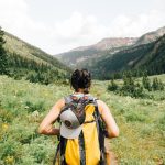 person carrying yellow and black backpack walking between green plants